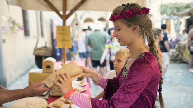 A girl dressed in a vibrant purple medieval gown and floral headband is buying traditional sweets wrapped in paper at a market stall during the medieval festival in Volterra, Italy. With a young brother by her side, she engages with a vendor while the colorful, festive atmosphere bustles in the background. The table is filled with handmade treats.
Shot with Nikon D850