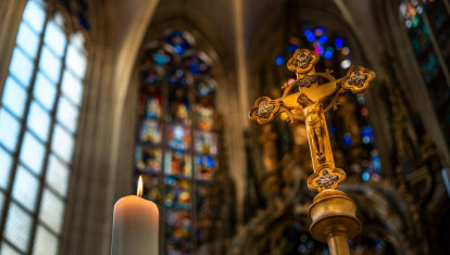 Close-up of ornate golden crucifix in a church interior, with blurred stained glass windows and vaulted ceiling in the background.