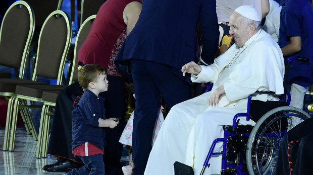 A two-year-old child playing onstage stares at Pope Francis, seated in a wheelchair following knee treatment, during an audience as part of the Festival of Families - 10th World Meeting of Families, on June 22, 2022 at Paul-VI hall in The Vatican. (Photo by Filippo MONTEFORTE / AFP)