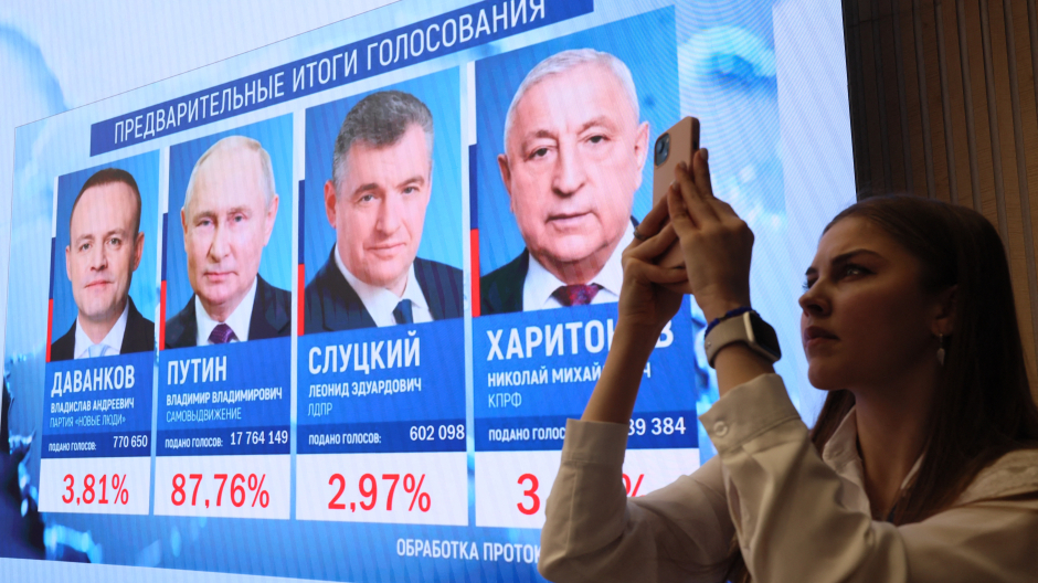Una mujer toma una fotografía con su teléfono móvil frente a una pantalla con los resultados preliminares de las elecciones presidenciales rusas en la Comisión Electoral Central en Moscú