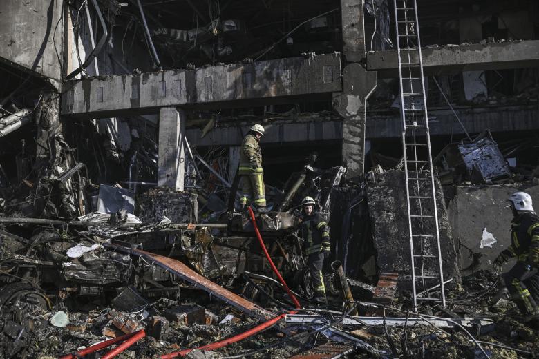 Los bomberos se paran en un vehículo militar blindado destruido en el centro comercial Retroville después de un ataque ruso en el noroeste de la capital, Kiev