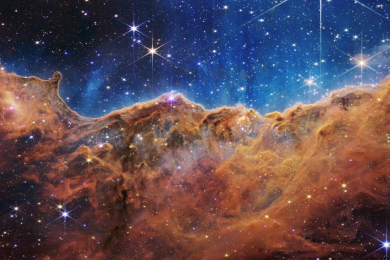 Este paisaje de "montañas" y "valles" salpicado de estrellas brillantes es en realidad el borde de una joven región de formación estelar cercana llamada NGC 3324 en la Nebulosa Carina. Capturada en luz infrarroja por el nuevo Telescopio Espacial James Webb de la NASA, esta imagen revela por primera vez áreas previamente invisibles de nacimiento de estrellas.