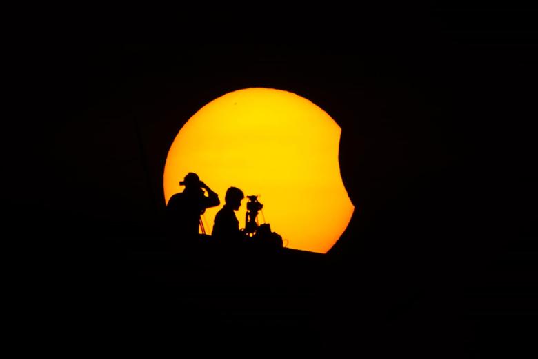 Dos personas se encuentra en lo alto de un edificio mientras la luna cubre parcialmente el sol durante un eclipse solar parcial, en Marina Beach, en Chennai, India.