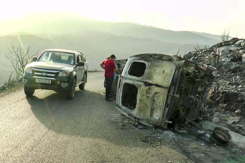 Varias personas inspeccionan vehículos quemados en una carretera de montaña después de los incendios forestales en los bosques de Bejaia, en el norte de Argelia