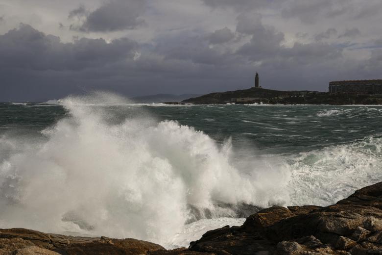 La Coruña sigue en alerta roja por la fuerza del viento y las olas. En la imagen una ola y al fondo la Torre de Hércules