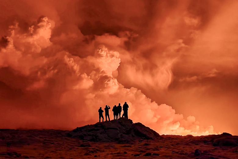 Unos residentes locales observan cómo se eleva el humo mientras la lava tiñe el cielo nocturno de naranja debido a la erupción volcánica