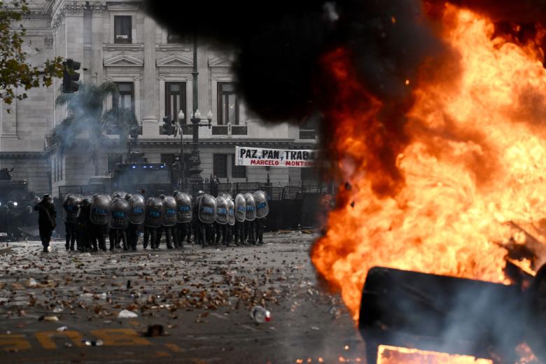 Agentes de la policía antidisturbios hacen guardia detrás de un auto de la estación de radio Cadena 3 en llamas durante una violenta protesta frente al Congreso Nacional en Buenos Aires