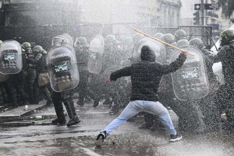 Un manifestante se enfrenta a la policía antidisturbios con un trozo de madera mientras lo rocían con un cañón de agua