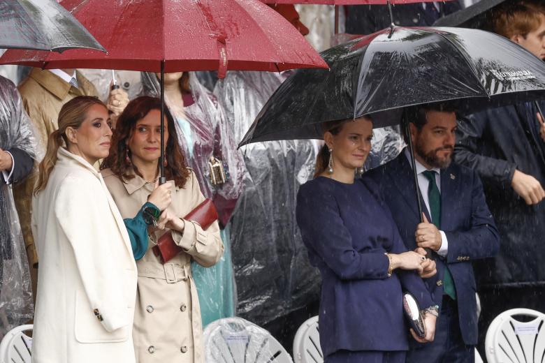 La presidenta de la Junta de Extremadura, María Guardiola, la presidenta de la Comunidad de Madrid, Isabel Díaz Ayuso; la presidenta de Baleares, Marga Prohens (2d), y su marido Javier Bonet (d), se protegen de la lluvia durante el desfile