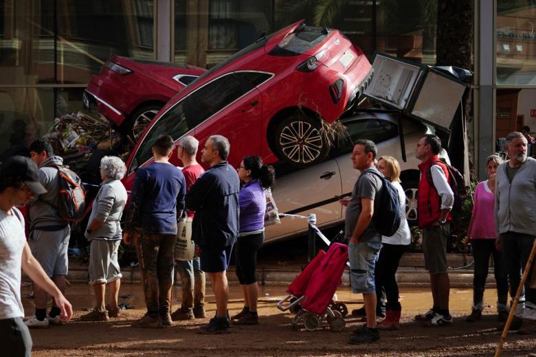 La gente hace cola para recibir suministros cerca de una montaña de coches destrozados por la DANA