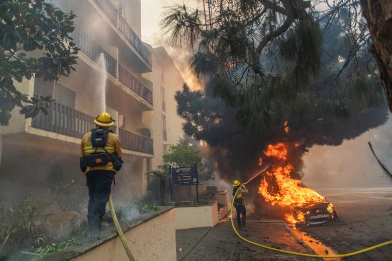 Los bomberos rocían agua hacia un automóvil y un complejo de edificios mientras las llamas del incendio Palisades ardían en Sunset Boulevard