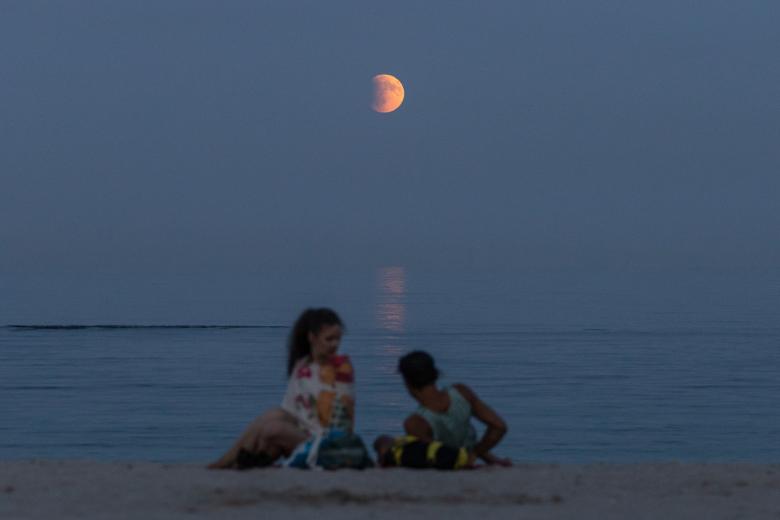 Los bañistas descansan en la orilla del Mar Negro con la luna llena, también conocida como 'Luna de Sangre', al fondo durante una fase de eclipse total en Odesa, Ucrania