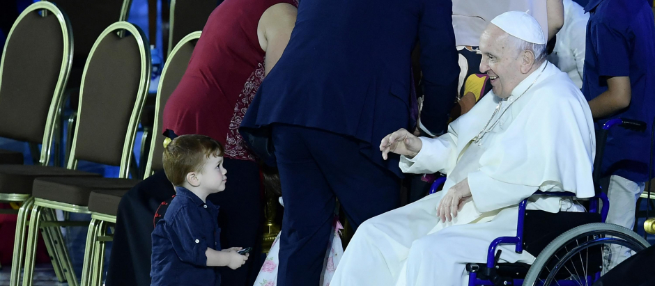 A two-year-old child playing onstage stares at Pope Francis, seated in a wheelchair following knee treatment, during an audience as part of the Festival of Families - 10th World Meeting of Families, on June 22, 2022 at Paul-VI hall in The Vatican. (Photo by Filippo MONTEFORTE / AFP)