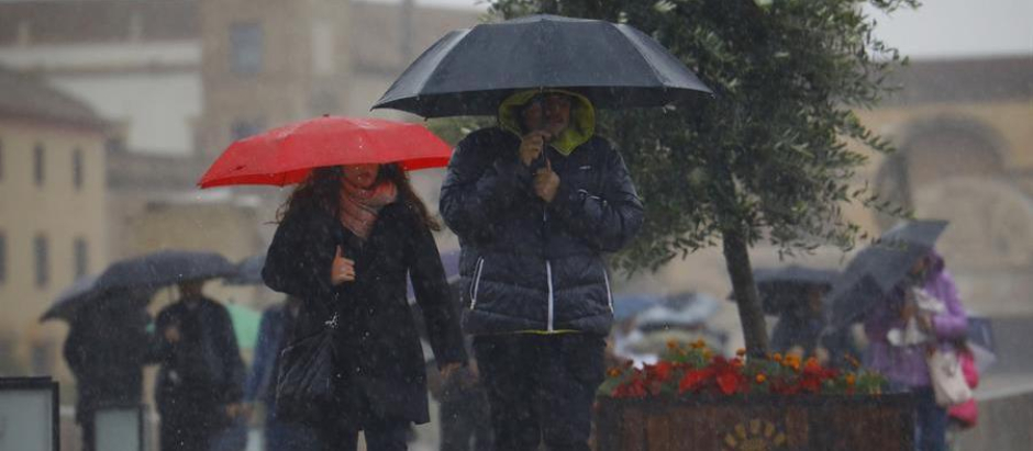 Unas personas se protegen con paraguas de la lluvia mientras caminan por el puente romano de Córdoba en una jornada en la que la ciudad se encuentra en aviso amarillo por lluvias