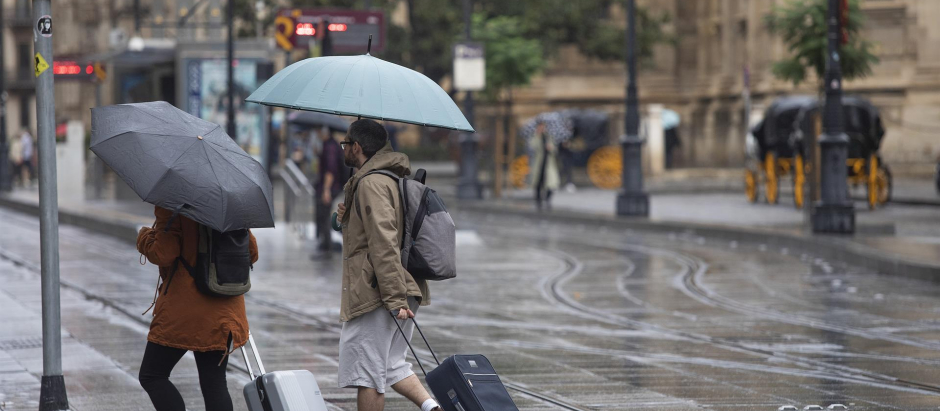 Una pareja con maletas se protegen de la lluvia bajo sus paraguas, en Sevilla (Andalucía, España)