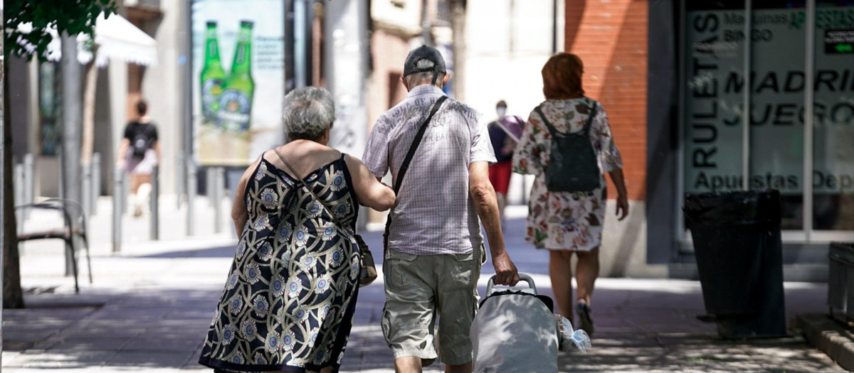 Una pareja de ancianos camina por la calle con un carro de compra.