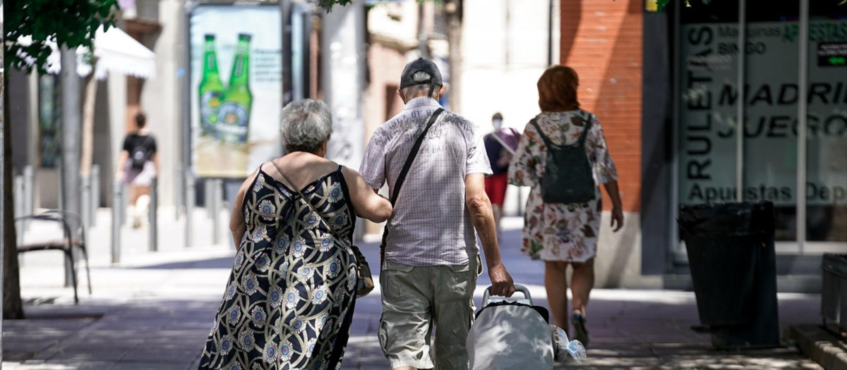 Una pareja de ancianos camina por la calle con un carro de compra.