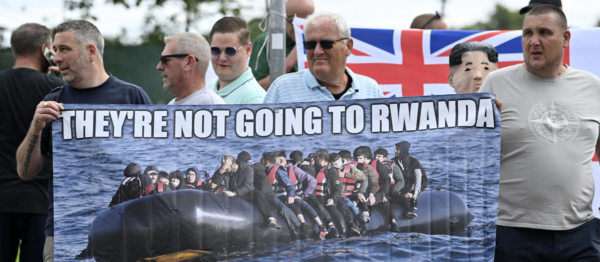 Protesters hold a banner depicting migrants crossing the Channel on a dinghy reading "They're not going to Rwanda, they're coming to Rushmoor" during a 'Enough is Enough' demonstration called by far-right activists near a hotel housing asylum seekers in Aldershot on August 4, 2024. Far-right protesters clashed with British police during tense rallies as unrest linked to disinformation about a mass stabbing that killed three young girls spread across the UK. (Photo by JUSTIN TALLIS / AFP)