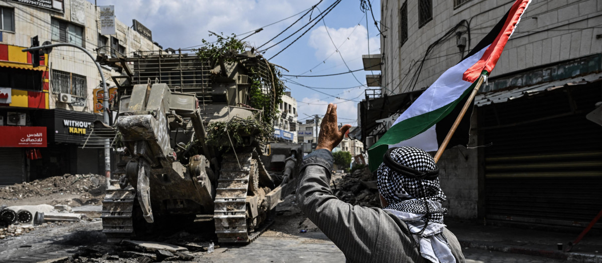 Un activista palestino sostiene una bandera nacional frente a una excavadora que daña una calle durante una redada israelí en el centro de Yenín, en Cisjordania