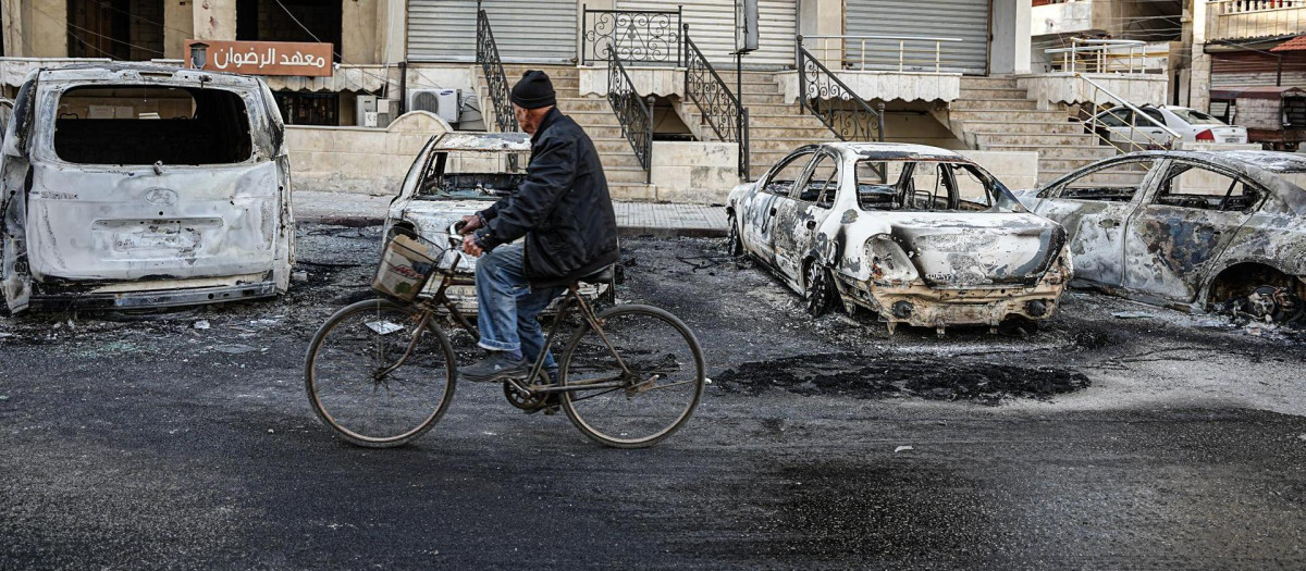 Un hombre sirio pasa en bicicleta junto a vehículos dañados tras la violencia ocurrida en los días anteriores durante los enfrentamientos entre fuerzas gubernamentales y partidarios del antiguo régimen sirio, en la ciudad de Jableh, en Latakia, Siria