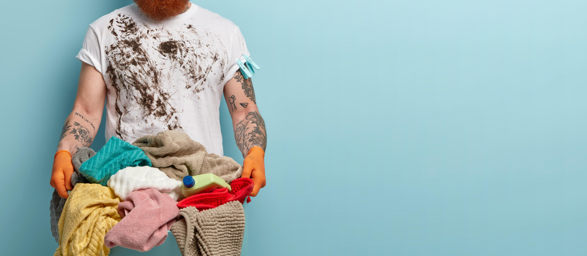 Puzzled dirty housekeeper wears casual white t shirt, holds basket with linen, does laundry during weekend, looks surprisingly at camera, has red messy hair, poses over blue studio wall. Household