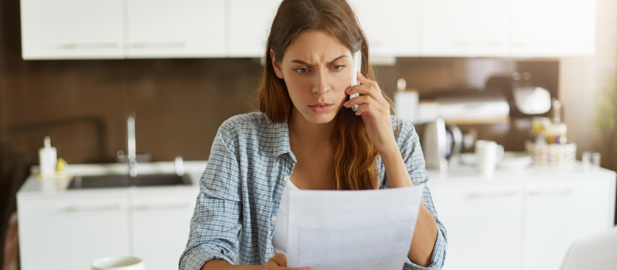 Candid shot of worried young European female dressed casually sitting at kitchen table, holding paper sheet and talking to house and utilities service representative about miscalculation in bills
