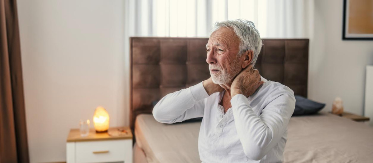 Senior adult man with gray hair and beard suffering from neck pain and stiffness, sitting on his bed in pajamas, feeling discomfort in the morning at home