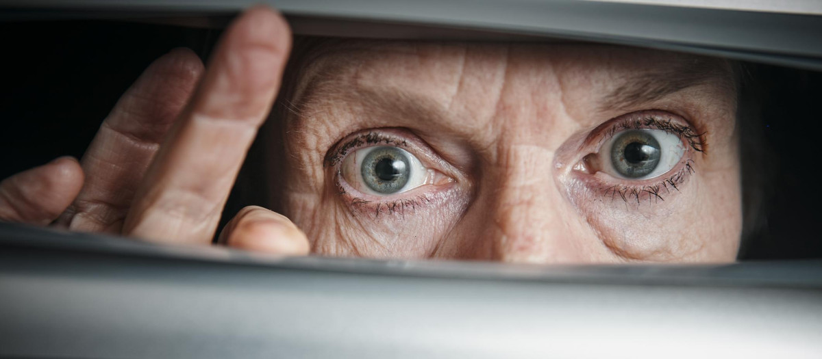 A senior woman peeps fearfully through venetian blinds