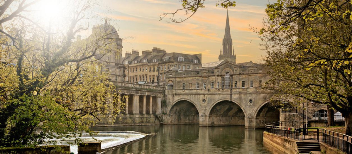 Historic Pulteney Bridge in Bath, England, UK at dusk with sun and colorful skies.