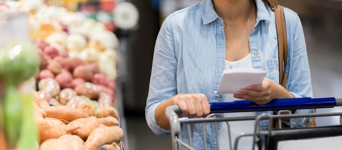 An unrecognizable woman pushes her cart past the potatoes in the grocery store.  She holds a shopping list.