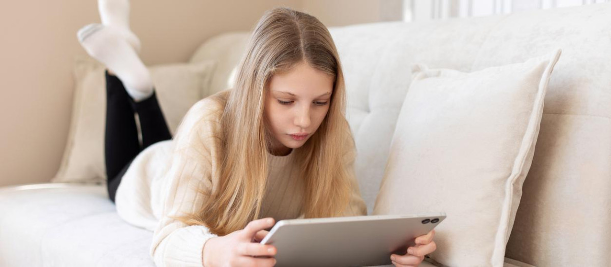 Teen girl lying on couch with tablet. Young girl lying comfortably on a sofa using a tablet, focused and relaxed in a cozy, well-lit living room setting