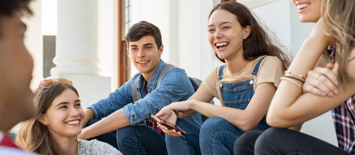 Group of happy young friends sitting in college campus and talking. Cheerful group of  smiling girls and guys feeling relaxed after university exam. Excited millenials laughing and having fun outdoor.