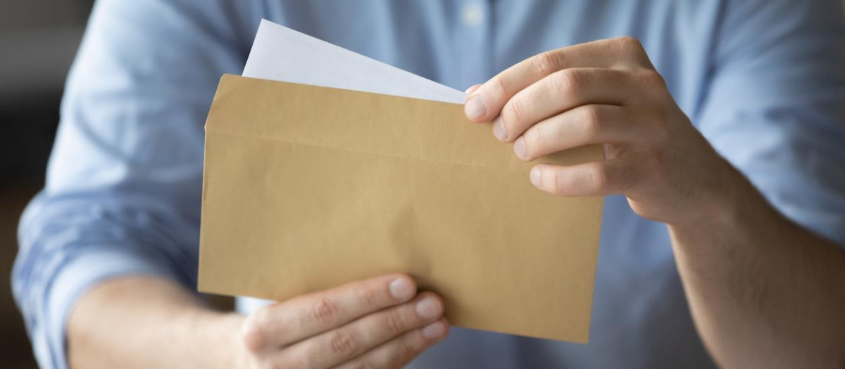 Businessman sitting at workplace desk holding yellow envelope take out received paper letter notice sheet, close up image. Business correspondence, corporate news, notification or invitation concept