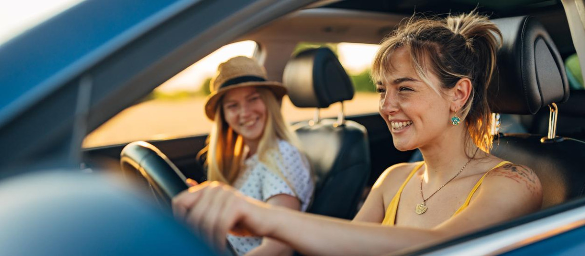 A smiling woman in casual, summer clothes is traveling on a summer vacation with her friend by car. She drives a car and enjoys the company of her best friend