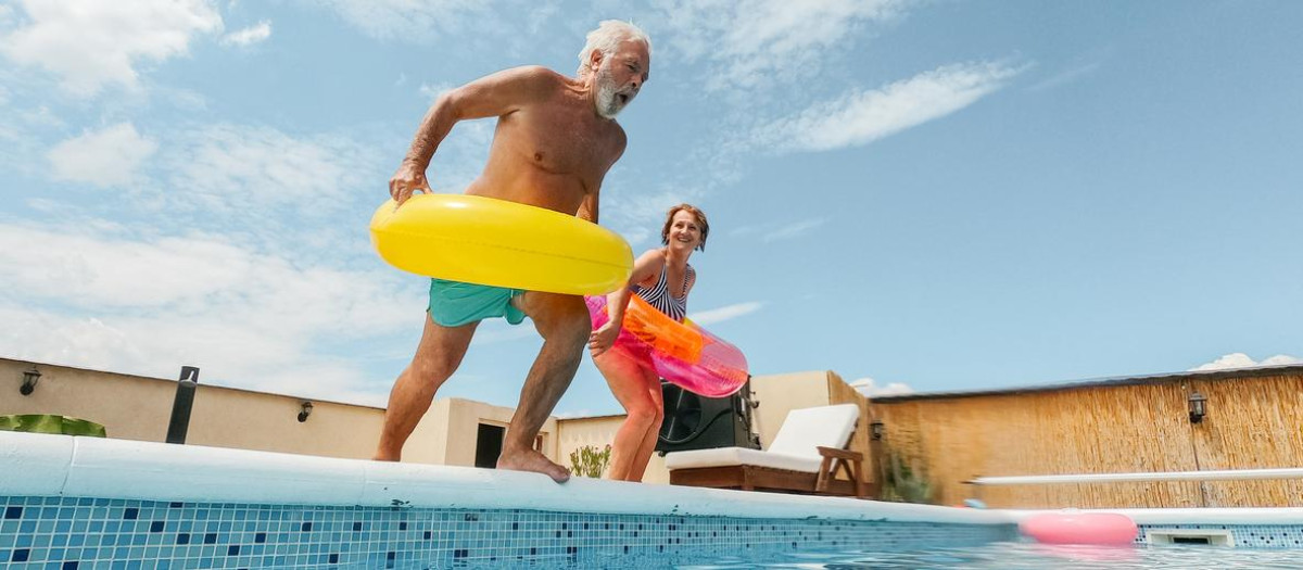 Photo of a senior couple refreshing on a hot summer day in 
 swimming pool; they are jumping into the pool while wearing inflatable rings