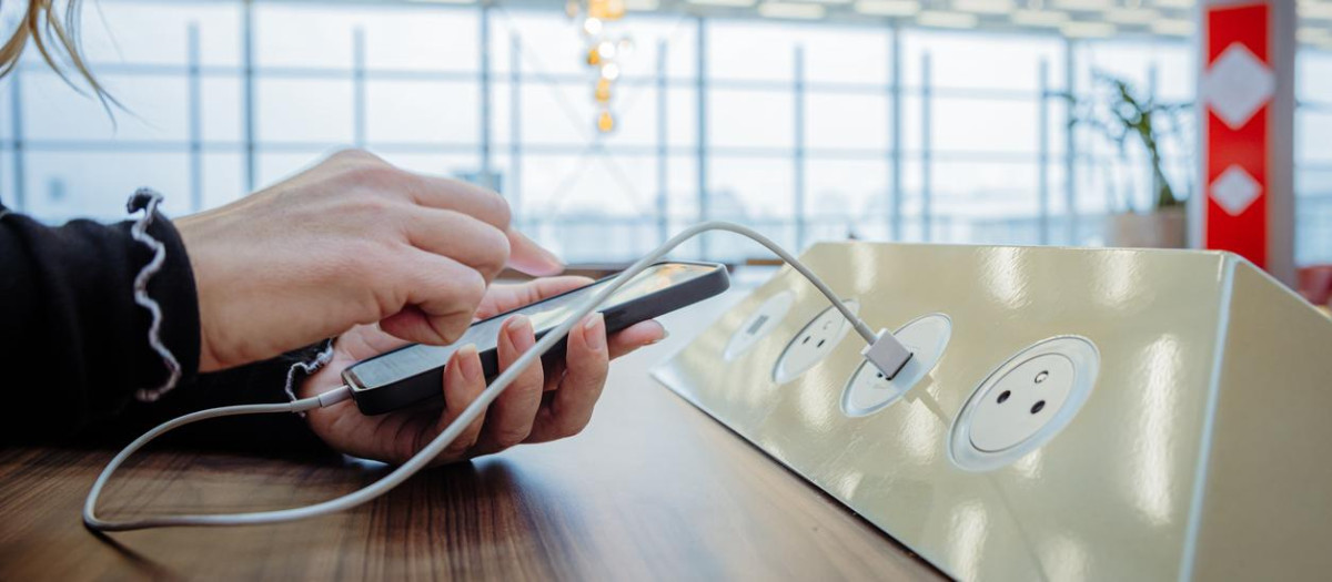Close-up of a woman with blond hair, wearing a black pullover, using her smartphone while charging it via a white USB cable on a public charging station installed on a wooden table inside an airport terminal, window of the terminal in the background, focus on forefront, horizontal