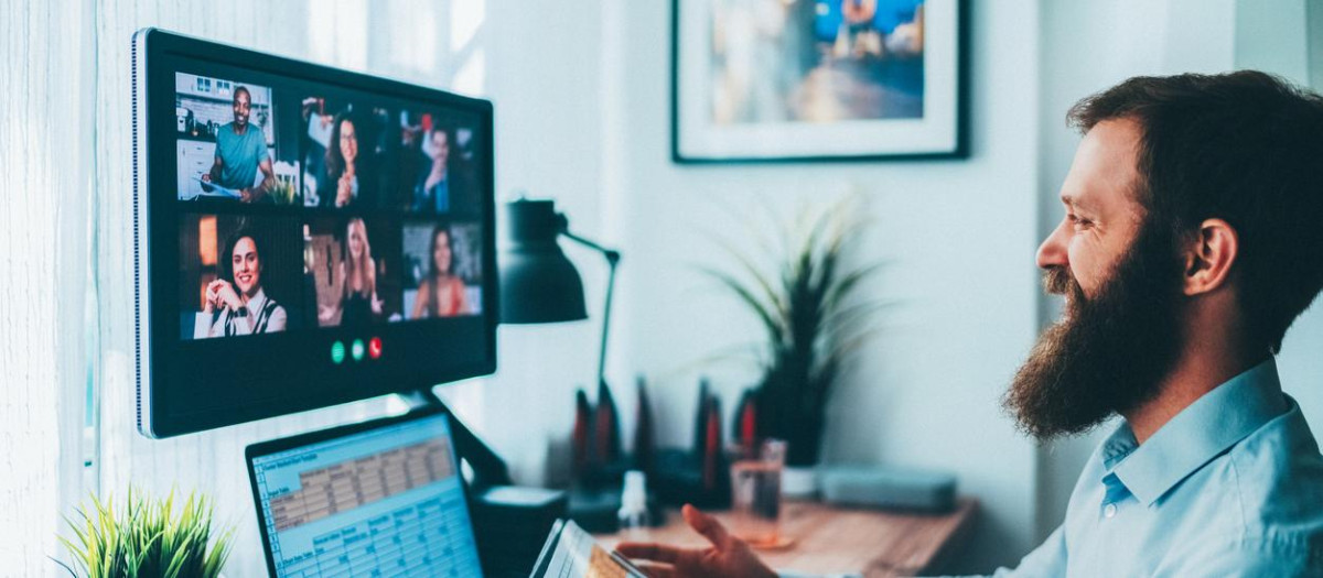 A professional works from a modern home office, participating in a remote team meeting via video call on a large screen while analyzing graphs and data on a digital tablet and laptop. The workspace is neatly organized with technology devices, plants, and office supplies, illustrating digital collaboration, productivity, and efficient remote work.