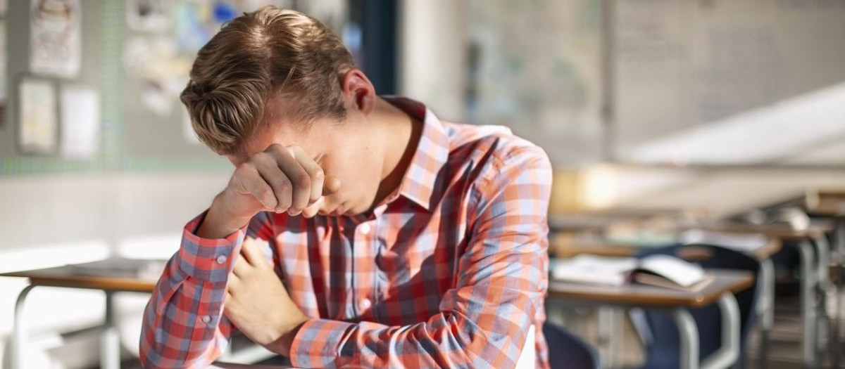 Stressed male student sitting with book at desk. Teenage boy is studying alone in classroom. He is at high school.