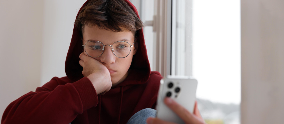 Loneliness concept. Sad teenage boy using smartphone near window indoors