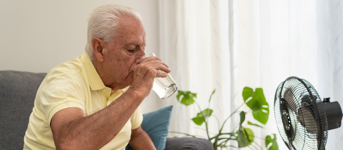 Senior man refreshing himself drinking water and using an electric fan during a hot summer at home