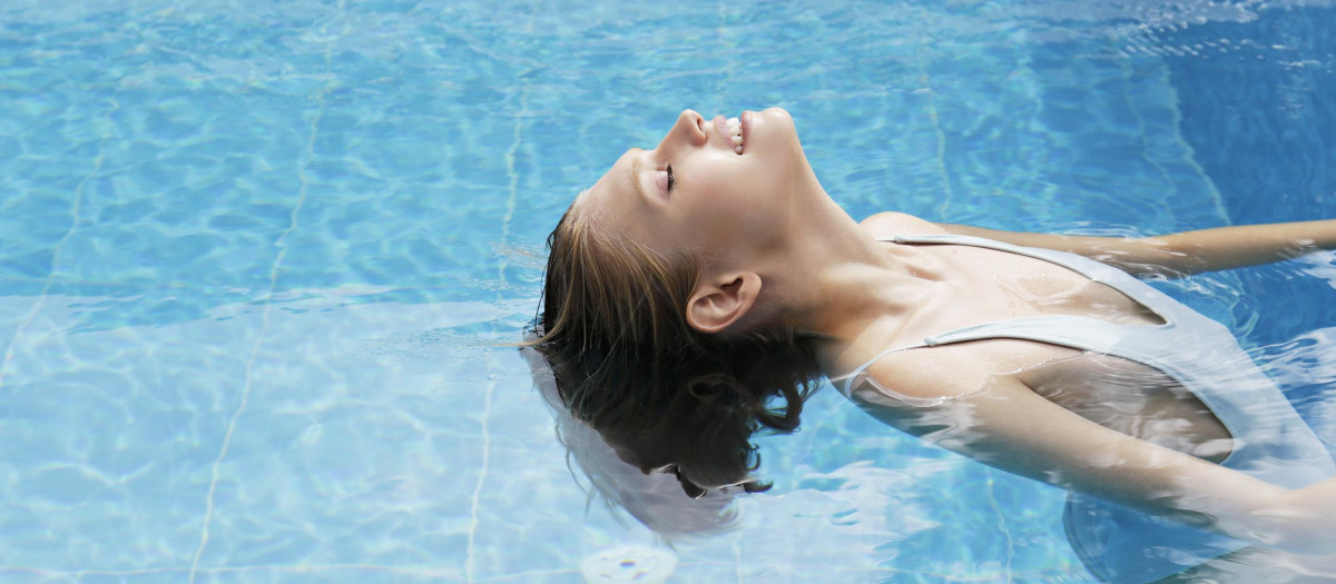 Fit young woman wearing blue one piece bathing suit emerging from the water. Female model with wet blonde hair cooling down in the pool on a hot summer day. Background, copy space, close up.