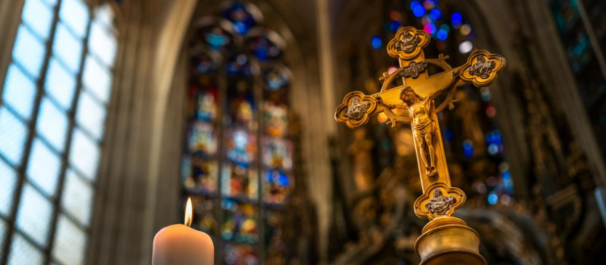 Close-up of ornate golden crucifix in a church interior, with blurred stained glass windows and vaulted ceiling in the background.