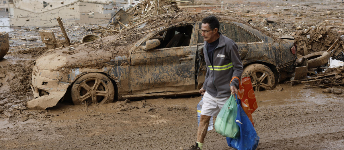 Un hombre con productos de primera necesidad pasa ante una zona anegada de barro en Paiporta