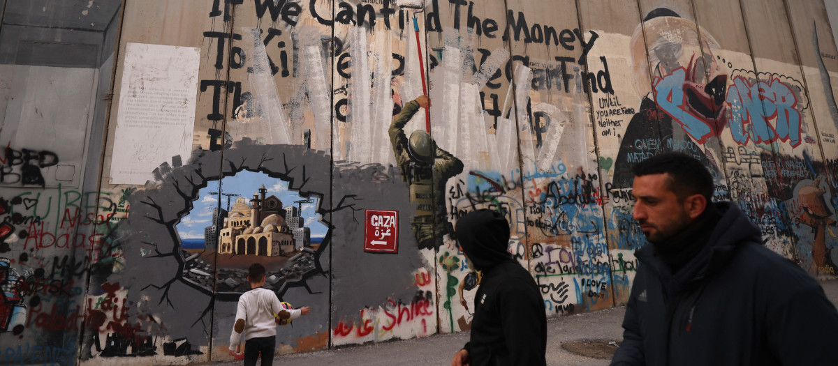Un niño juega mientras unos hombres pasan junto a un grafiti que representa la reconstrucción de Gaza, en una sección de la barrera de separación de Israel, en Belén, en la Cisjordania