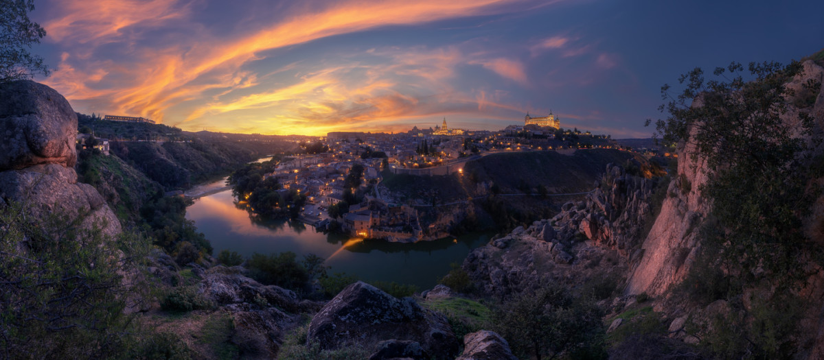 Sunset over the historic city of Toledo seen from Mirador del Valle. Dramatic clouds, river reflection, and golden evening light illuminate the skyline.