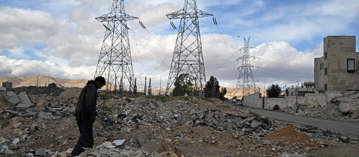Un hombre camina entre los escombros de edificios con torres de alta tensión como telón de fondo cerca de Damasco