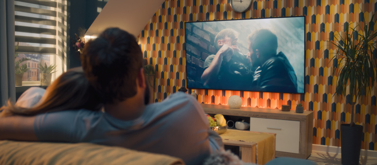 Couple on sofa in living room, watching action movie on TV or criminal blockbuster on streaming service, talking and discussing acting, resting at home on weekend. Home theater in modern apartment.