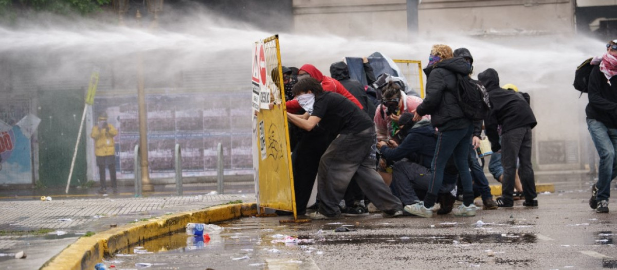 Protesters shield themselves with an improvised barrier as police spray them with water from a water cannon during a university march in Buenos Aires, Argentina, on October 15, 2026. (Photo by Guillermo Castro/NurPhoto) (Photo by Guillermo Castro / NurPhoto via AFP)