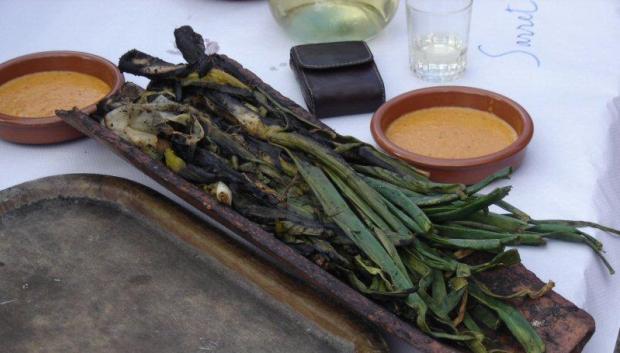 Una teja llena de 'calçots' junto a dos platos con salsa romesco