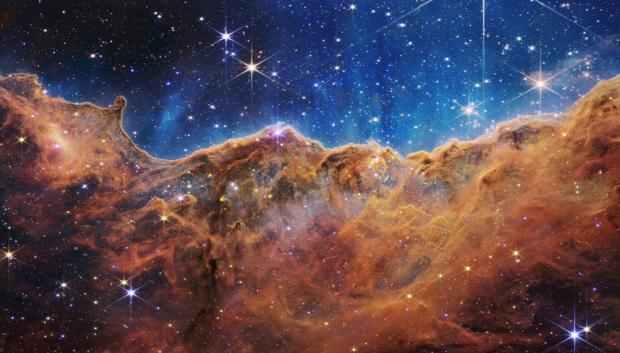Este paisaje de "montañas" y "valles" salpicado de estrellas brillantes es en realidad el borde de una joven región de formación estelar cercana llamada NGC 3324 en la Nebulosa Carina. Capturada en luz infrarroja por el nuevo Telescopio Espacial James Webb de la NASA, esta imagen revela por primera vez áreas previamente invisibles de nacimiento de estrellas.